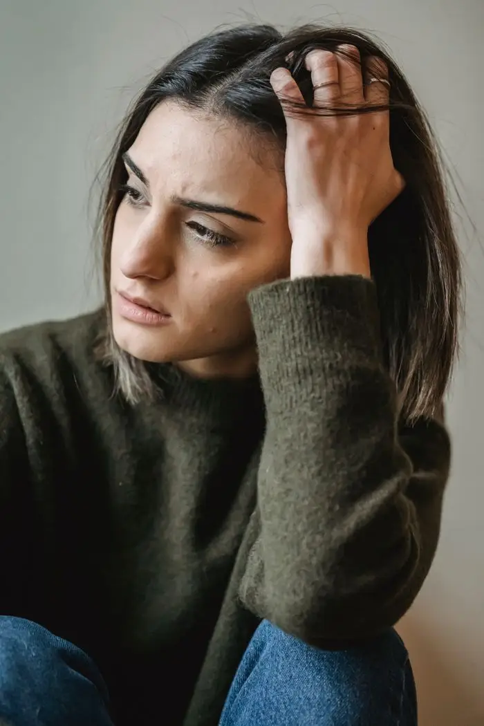 Thoughtful young ethnic woman in casual clothes touching dark hair and looking away while sitting near white wall